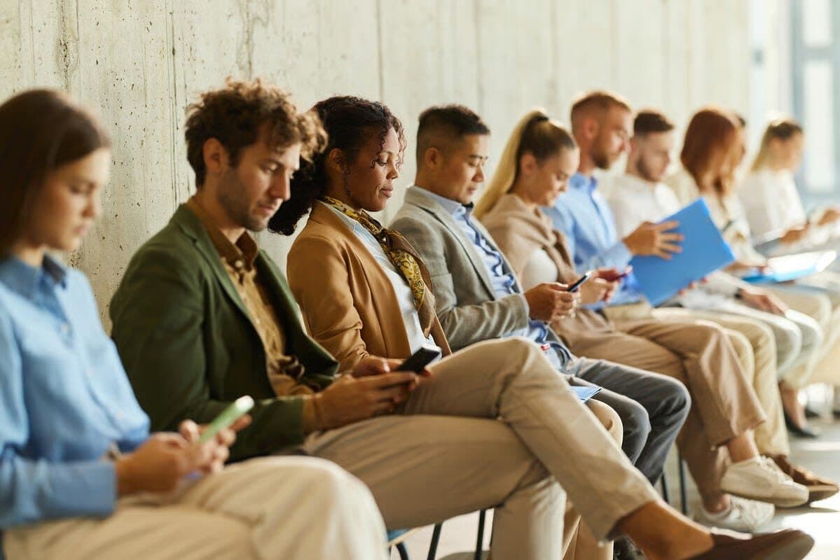 A row of people seated against a concrete wall, looking at their phones